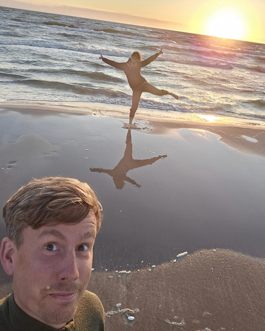 Aga balancing on one leg with her arms out at the edge of the sea at sunset, Thomas pulling a face in the foreground
