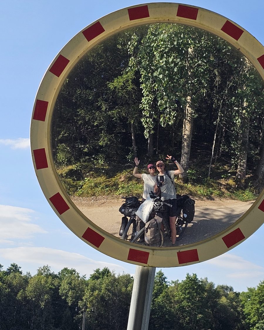 Thomas and Aga waving in a roadside traffic mirror, their loaded bikes in front of them on a gravel road