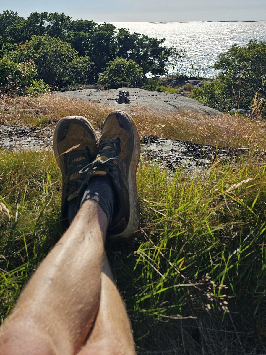 Crossed legs and worn trail shoes in the grass on a rock, the sea glittering beyond the trees