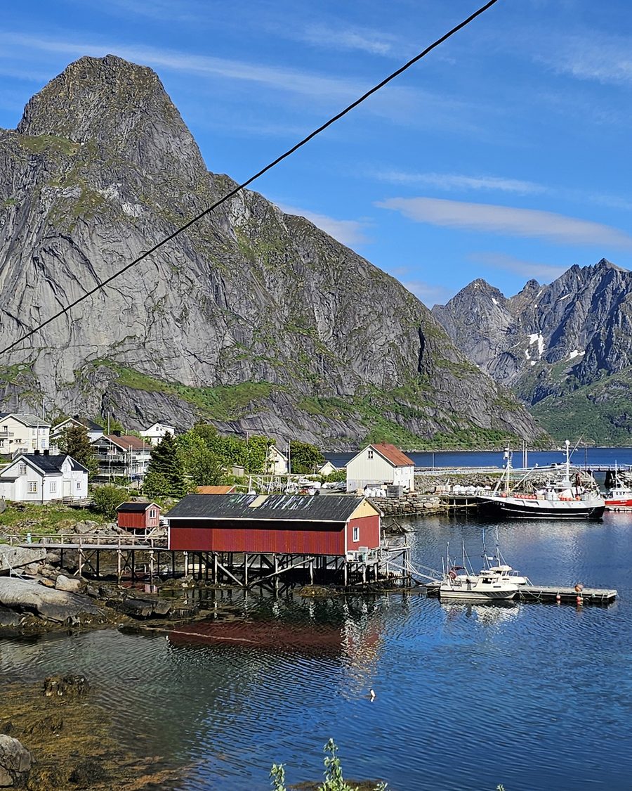 Reine: a red boathouse on stilts and fishing boats under a steep grey peak, a cable crossing the sky