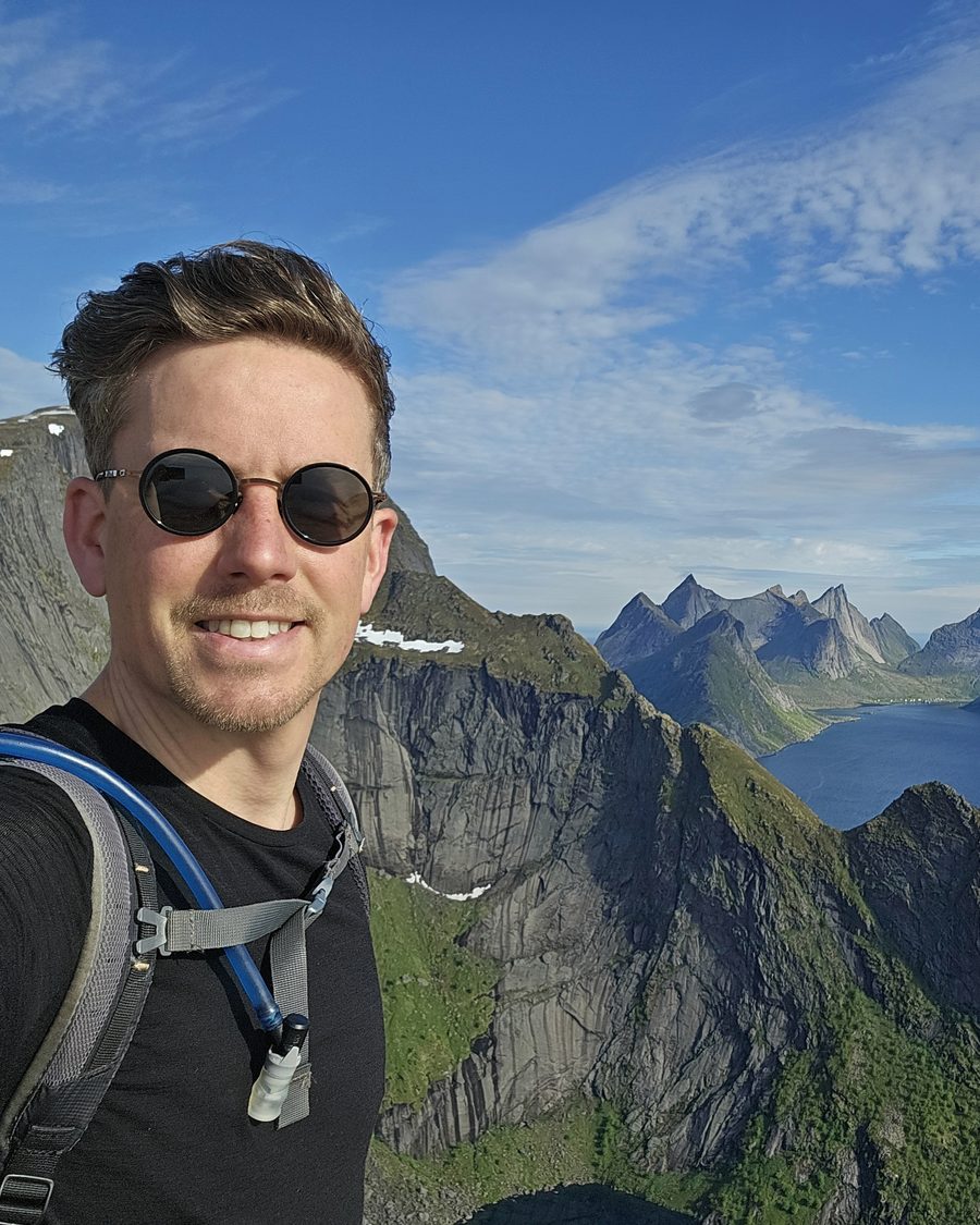 Thomas on top of Reinebringen, the fjord and jagged peaks behind him