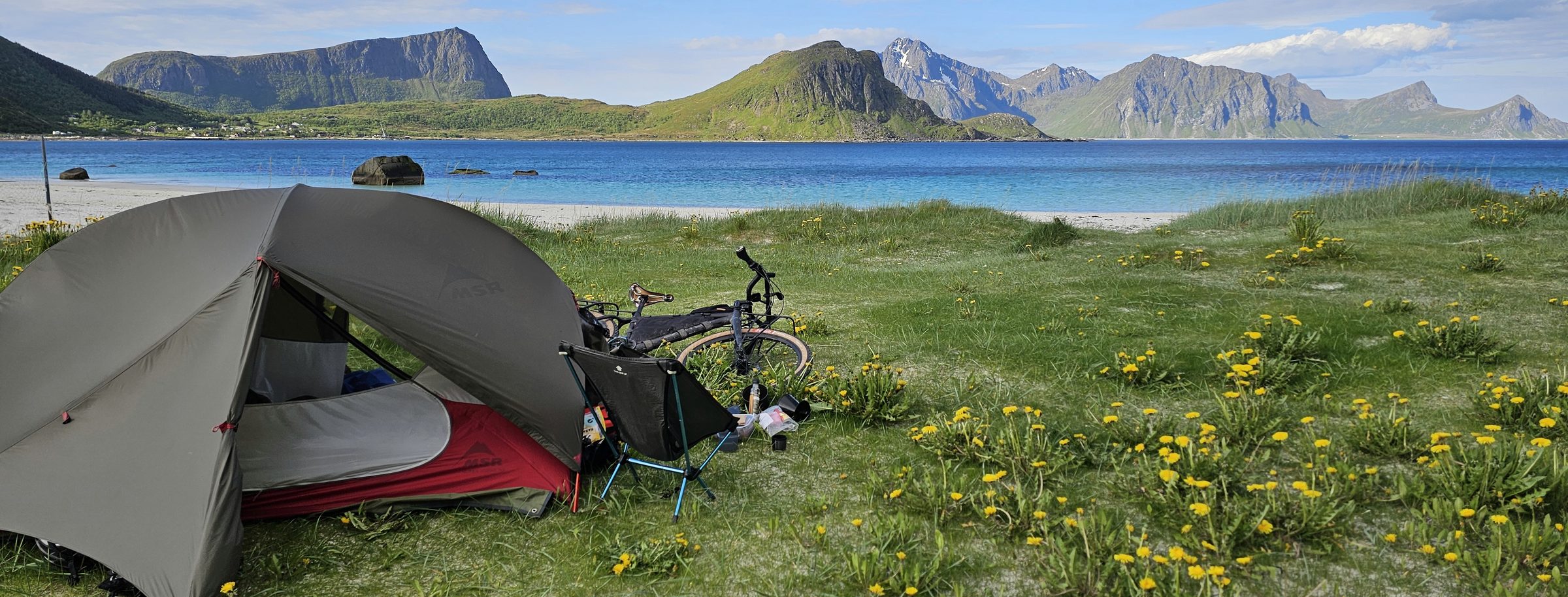 Tent, camp chair and bike in the dandelions above a white beach, turquoise water and mountains behind