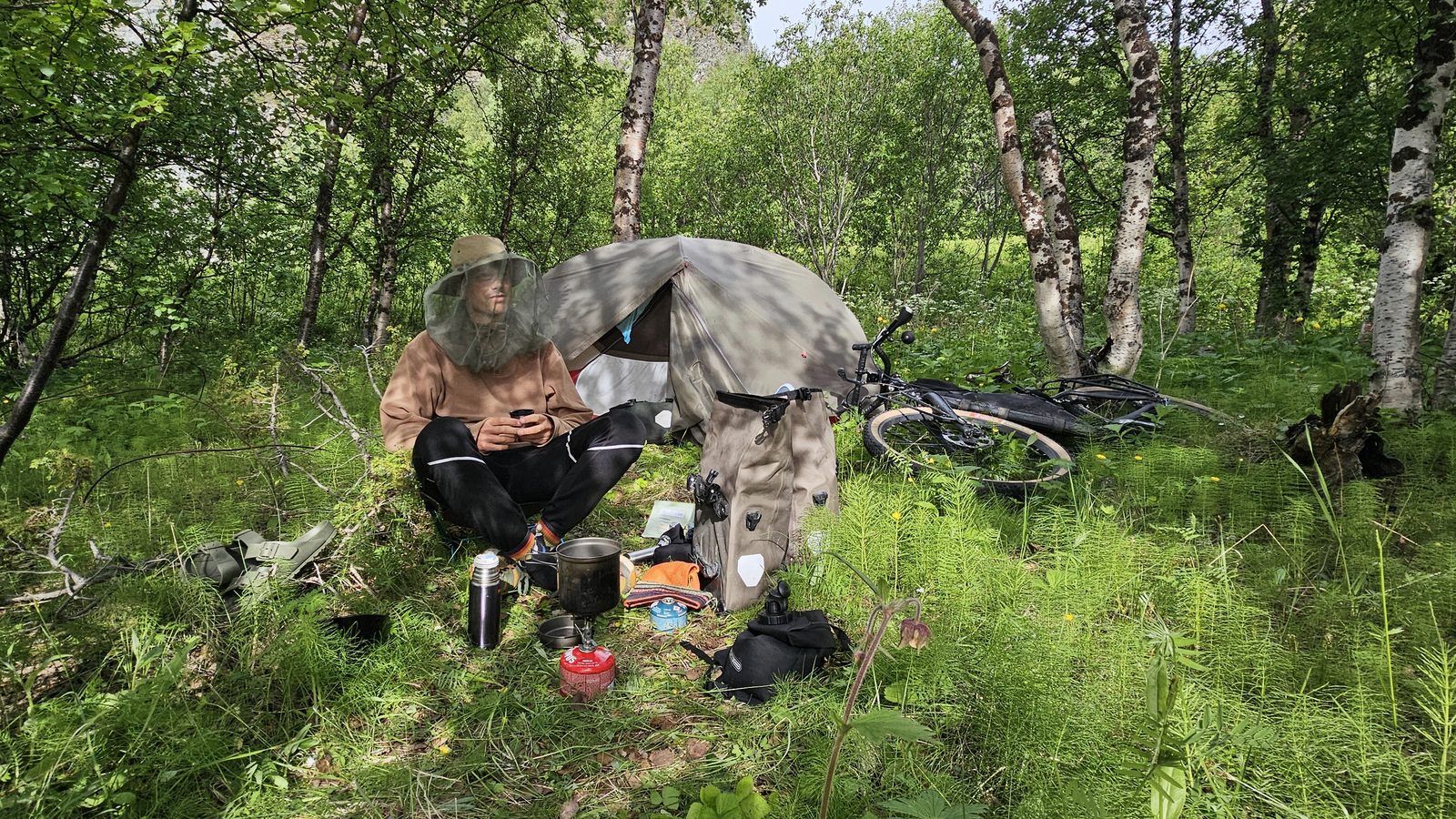 Thomas sitting in front of his tent in a birch wood, wearing a hat with a mosquito net, stove and thermos in the grass