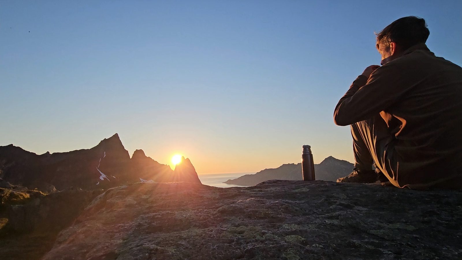 Thomas sitting on a rock on Hesten with a thermos, the midnight sun low behind jagged peaks