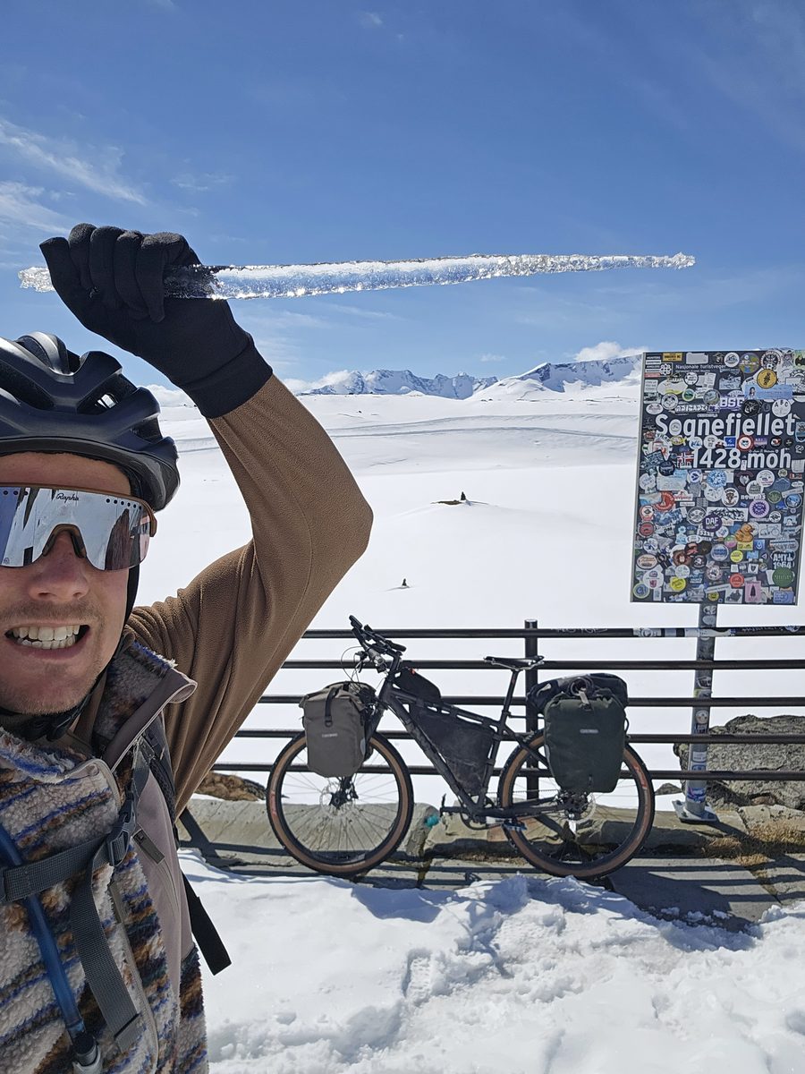 Thomas holding up an icicle next to the Sognefjellet summit sign, loaded bike against the railing, snow all around